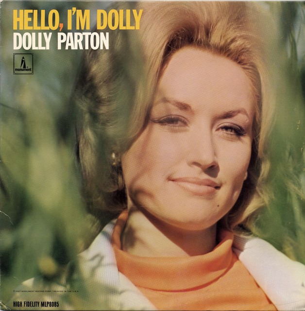 Dolly Parton on the Hello, I'm Dolly album cover, smiling in an orange turtleneck against blurred green foliage